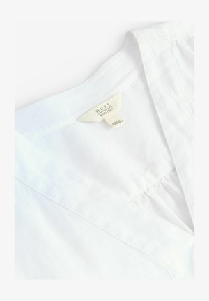 Baltas lina krekls ar V veida izgriezumu, ar uzrakstu "next 100% LINEN." Gluda tekstūra, viegls audums, vienkāršas šūšanas detaļas.