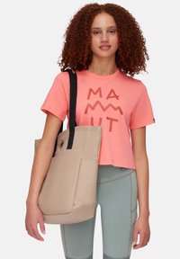 Sac fourre-tout beige avec une texture rembourrée et des poignées noires, tenu dans la main d'une personne, associé à un t-shirt rose avec des lettres en bloc marron.