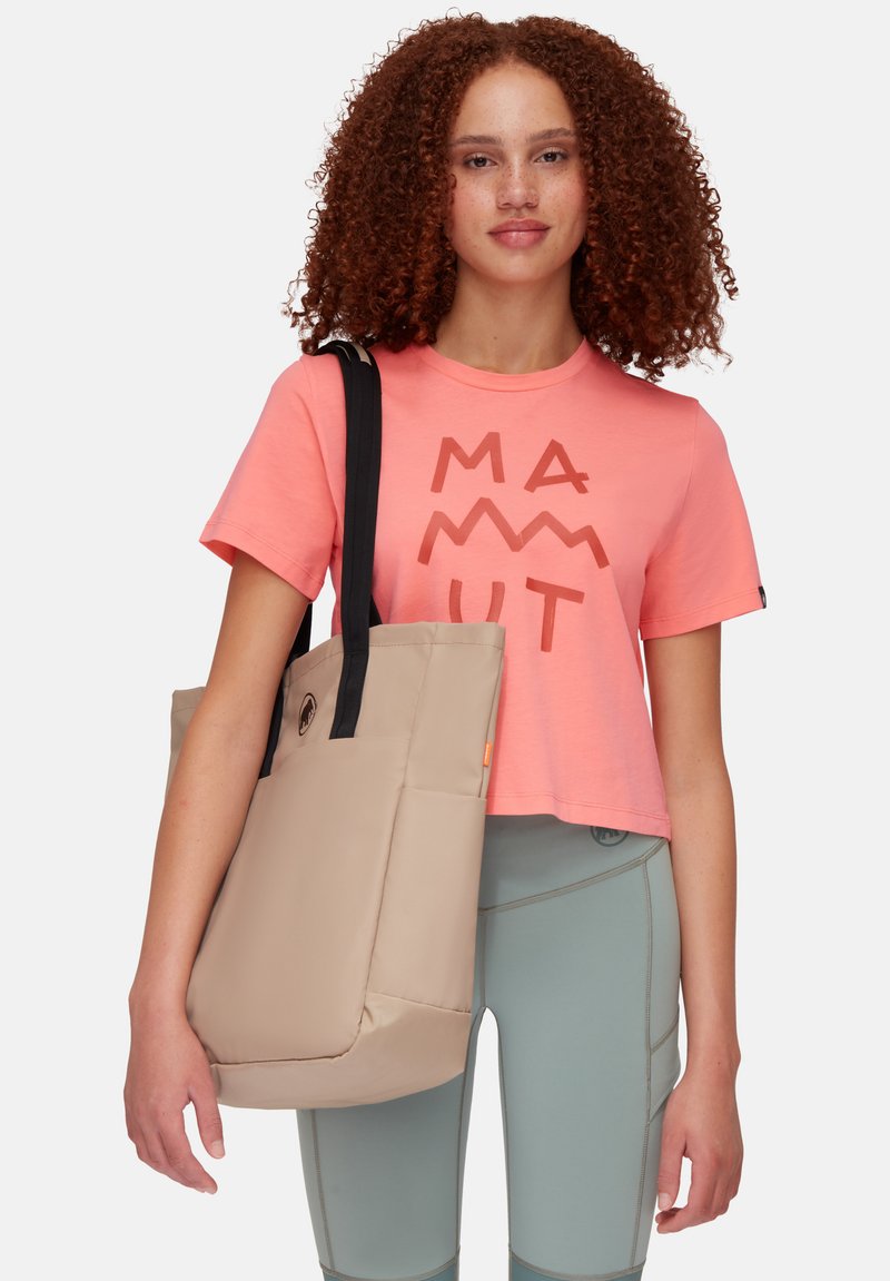 Sac fourre-tout beige avec une texture rembourrée et des poignées noires, tenu dans la main d'une personne, associé à un t-shirt rose avec des lettres en bloc marron.