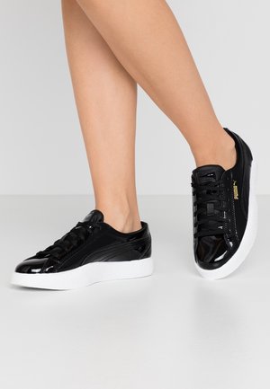 Zapatillas de charol negras con una suela blanca texturizada, cuentan con un diseño de cordones y un acento de logo dorado en el lateral. Superficie suave y silueta moderna.