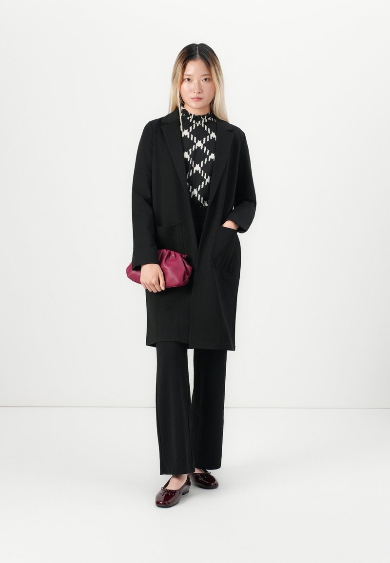 Frau steht in einem schwarzen langen Mantel, gemustertem schwarz-weißem Oberteil, schwarzen Hosen, burgunderfarbenen Ballerinas und hält eine burgunderfarbene Clutch-Tasche.