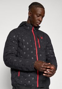 Black quilted jacket with a hood, featuring a subtle pattern, red zipper, and logo. Smooth texture, fitted design, and elastic cuffs.