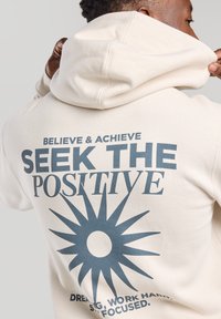 Sudadera de un beige claro con un bolsillo frontal, que presenta un gran gráfico en la parte posterior: un diseño de sol con el texto "BUSCA LO POSITIVO" en azul oscuro.