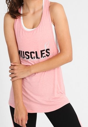 Kobieta w różowym topie z tekstem "MUSCLES" i czarnych legginsach, trzyma jedną rękę na ciele.