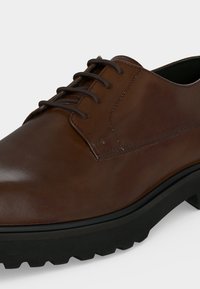 Καφέ δερμάτινο επίσημο παπούτσι με μαύρα κορδόνια και παχύ μαύρο καουτσούκ σόλα με βαθιά πέλματα για ενισχυμένη πρόσφυση.