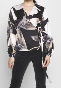Zwarte wikkelblouse met een bloemenpatroon in beige, grijs en wit. Lange mouwen met elastische manchetten en een strikdetail in de taille. Gladde stof.