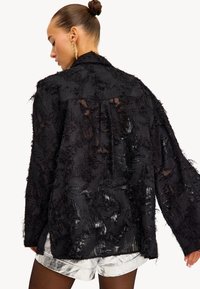 Zwarte doorschijnende shirt met lange mouwen, voorzien van textuur en franjes, relaxte pasvorm en iets verlengde achterkant. Gepaard met glanzende zilveren shorts.