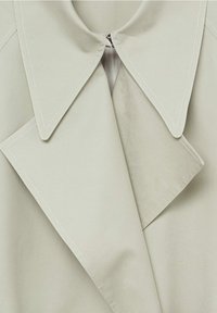 Lys grå frakke med en stor krave, rene linjer og synlige stikninger langs kanterne. Lynlås lukning foran. Blød stoftekstur.