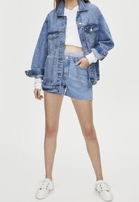 Nainen, joka on pukeutunut ylipirteään siniseen farkkutakkiin, valkoiseen lyhyeen toppiin, korkeavyötäröisiin farkkushortseihin, joissa on ketjuyksityiskohtia, ja valkoisiin lenkkareihin, seisoo käsi taskussa.