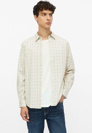 Junger Mann mit braunen lockigen Haaren trägt ein beiges gemustertes Hemd mit Knöpfen über einem weißen T-Shirt und blauen Jeans, steht mit einer Hand in der Tasche.