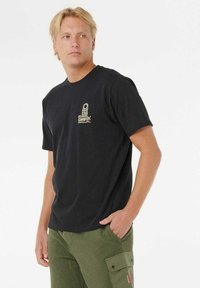 Sort bomulds T-shirt med rund hals og korte ærmer, med en guldbelagt logo detalje på den øverste venstre side. Bæres med grønne cargo bukser.