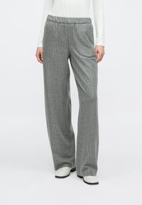 Femeie purtând pantaloni largi cu dungi gri, asortați cu pantofi albi cu vârf pătrat și un top cu mâneci lungi alb, stând pe un fundal alb.