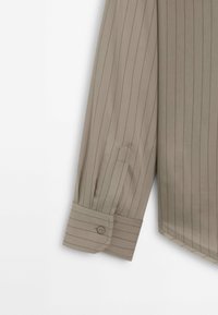 Chemise beige à manches longues avec fines rayures verticales, poignet boutonné et détail plissé sur la manche près du poignet.