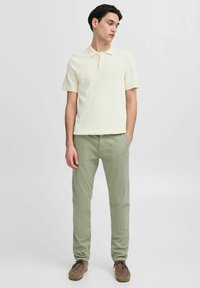Kremasta kratkodlana polo majica s teksturiranim pletenjem, uparena s svijetlozelenim chinos hlačama. Smeđe antilop cipele upotpunjuju outfit.