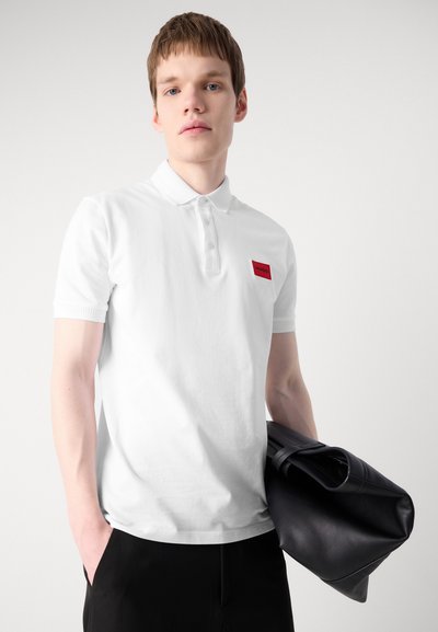 Polo shirt blanc à manches courtes et col côtelé. Équipé d'un patch logo rectangulaire rouge sur la poitrine gauche. Le modèle tient un sac en cuir noir.
