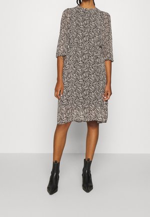 Kvinde iført en sort-hvid blomsterkjole, der går til knæene, med trekvartærmer og sorte ankelstøvler, stående foran en ensfarvet baggrund.