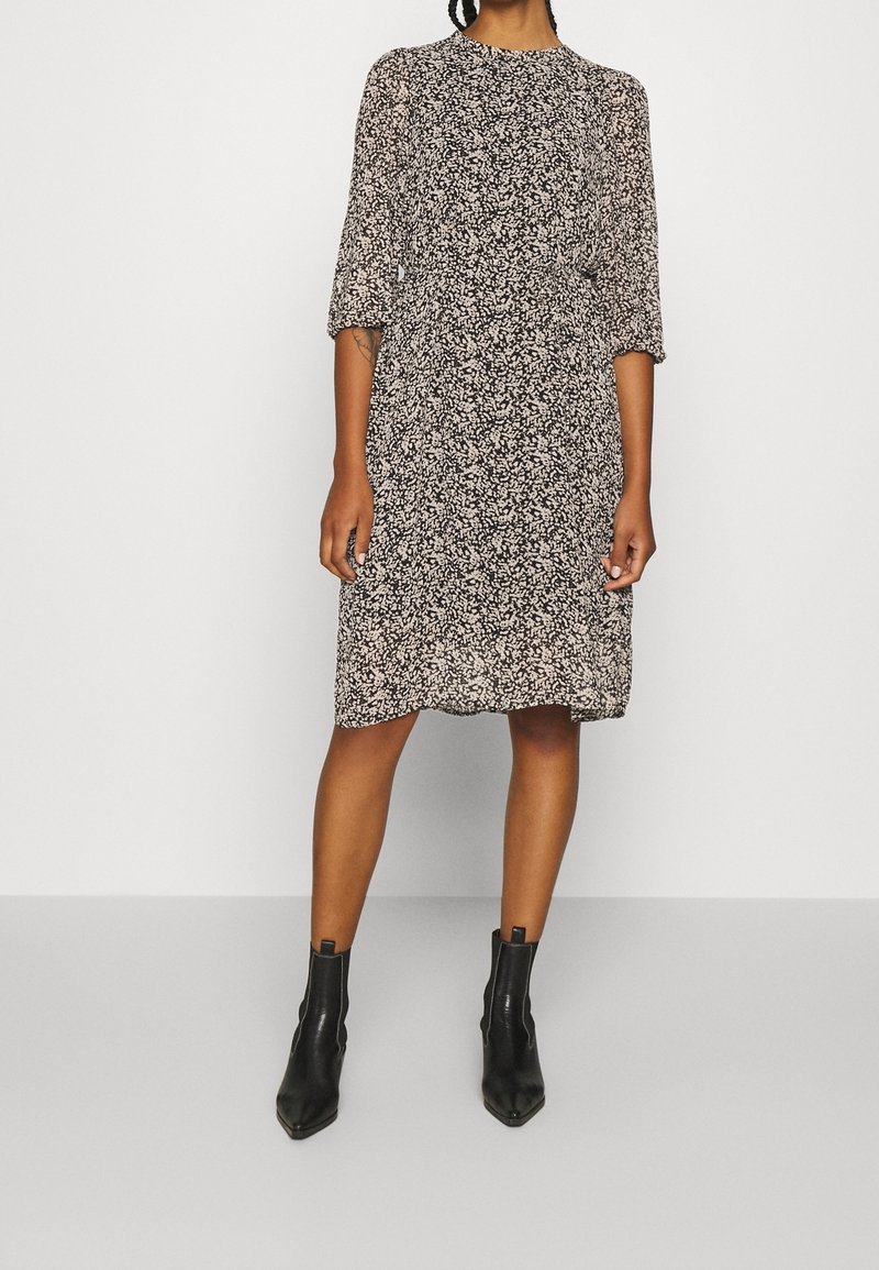 Femme portant une robe fleurie noire et blanche jusqu'aux genoux, avec des manches trois-quarts et des bottines noires, debout devant un fond uni.