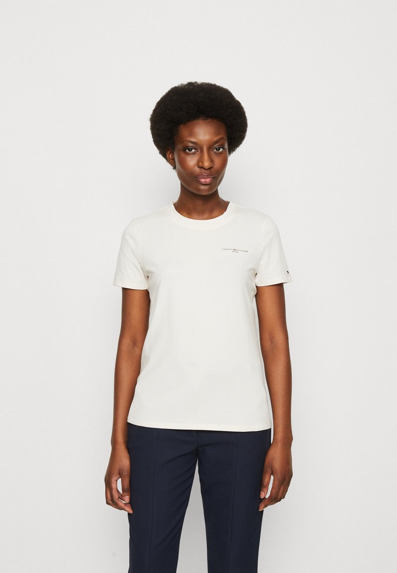T-shirt in cotone beige chiaro con maniche corte, colletto tondo e un piccolo logo stampato sul petto. Indossato con pantaloni blu navy.
