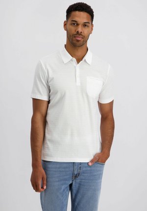 Homme portant un polo blanc à manches courtes avec une poche poitrine et un jean bleu, debout avec une main dans la poche devant un fond uni.