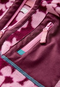 Polárdzseki részlet, amely virágmintát ábrázol rózsaszín és lila árnyalatokban, kontrasztos anyaggal, rózsaszín cipzárokkal és türkiz kiegészítő részletekkel.