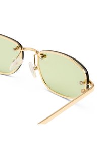 Sonnenbrille mit goldfarbenem Metallgestell, leicht grün getönten Gläsern und transparenten, verstellbaren Nasenpads auf weißem Hintergrund.