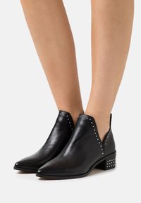 Bottines en cuir noir avec un bout pointu, des côtés découpés, des accents en clous argentés et un petit talon block. Texture lisse.
