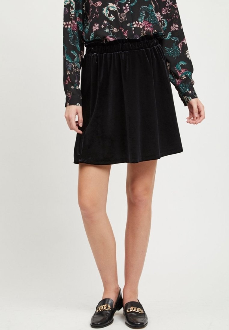 Jupe en velours noir avec taille élastique, longueur au-dessus du genou, texture lisse et coupe évasée, portée avec un chemisier à motifs et des mocassins.