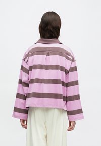 Άτομο που φορά μια χαλαρή, μακριά μπλούζα ροζ και καφέ με οριζόντιες ρίγες και φαρδύ γιακά, συνδυασμένη με κρέμα παντελόνια, εικονιζόμενο από πίσω.