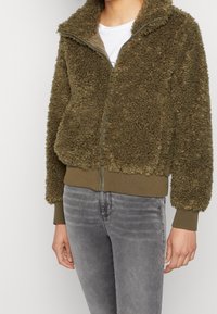 Olive green fleece jacket with a zip-up front, high collar, and ribbed cuffs and hem. Soft texture and cropped fit, paired with gray jeans.