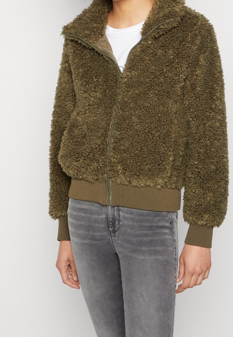 Olive green fleece jacket with a zip-up front, high collar, and ribbed cuffs and hem. Soft texture and cropped fit, paired with gray jeans.