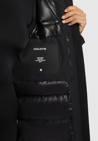 Veste matelassée noire avec une doublure intérieure brillante, arborant une étiquette portant la mention "KHUJOTM" et des détails de marque supplémentaires.