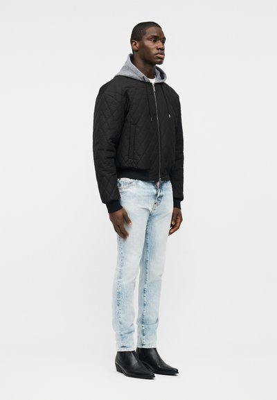 Homme portant une veste matelassée noire superposée à un sweat à capuche gris, un jean bleu clair et des bottines à bout pointu noires, se tenant légèrement de profil vers la droite.