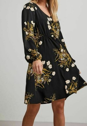 Zwarte jurk met lange mouwen met gele en witte bloemmotieven, V-hals, getailleerde taille en knielange zoom gedragen door een staand persoon.