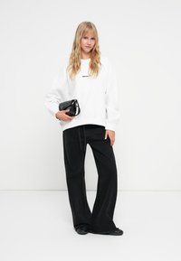 Biely oversize mikina s čiernym textom, spárovaná s čiernymi nohavicami s vysokým pásom a širokými nohavicami. Modelka drží malú čiernu kabelku.