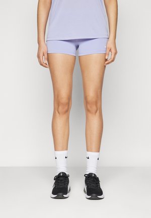 Person trägt hellviolette Sportshorts und Top, weiße Nike-Socken und schwarz-weiße Laufschuhe, steht vor einfarbigem Hintergrund.