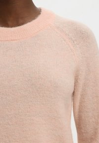Weicher rosa Strickpullover, getragen von einer Person, zeigt Rundhalsausschnitt und Details der Schulternaht in Nahaufnahme.
