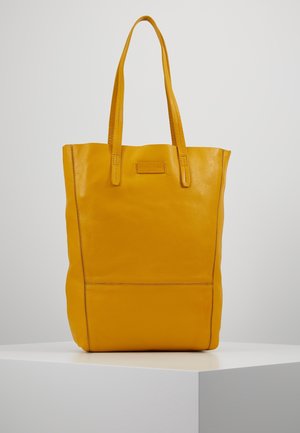 Żółta torba na ramię ze skóry z dwoma długimi uchwytami i przeszyciami, stojąca na białym postumencie na tle jasnego, jednolitego tła.