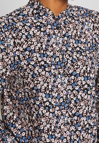 Camicetta nera floreale con colletto alto, caratterizzata da piccoli fiori bianchi, rosa e blu, e cinque bottoni scuri lungo la parte anteriore.