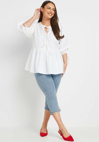 Valkoinen peplum-toppi pitkillä puff-hihoilla ja nauhakiinnityksellä kaula-aukossa, yhdistettynä vaaleansinisiin lyhyisiin farkkuihin ja punaisiin kärkitossuihin.