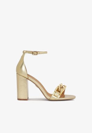 Sandal en or à talon haut, avec un talon bloc épais, une bride de cheville et un accent en chaîne métallique sur la bride avant. Fini lisse et brillant.