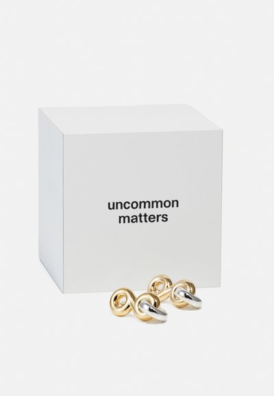 Boucles d'oreilles métalliques en or et argent avec des boucles entrelacées et une finition polie, présentées à côté d'une boîte blanche étiquetée "uncommon matters."