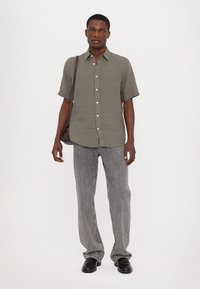 Korte mouwen olijfgroene shirt met knopen, gecombineerd met lichtgrijze gestructureerde wijde broek en zwarte schoenen. Bruine schoudertas.