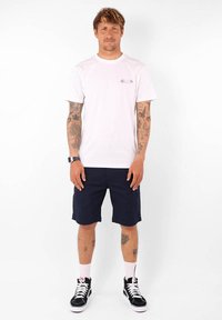 T-shirt en coton blanc avec un petit motif coloré, associé à un short bleu marine, des chaussettes blanches et des baskets noires avec des accents blancs.