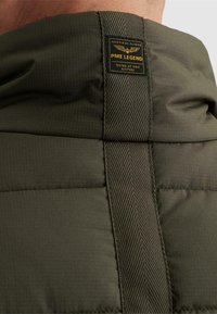 Gros plan du col d'une veste matelassée vert olive avec une sangle verticale texturée et une étiquette de marque PME Legend en noir et jaune.