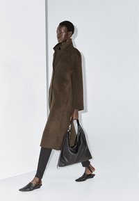 Lang brun uldfrakke med højt krave, båret sammen med sorte tætsiddende bukser og mules, og bærer en blød sort læder håndtaske.