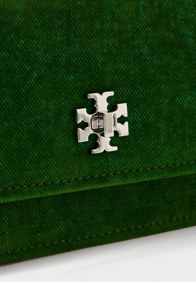 Srebrni kovinski logo emblem v obliki simetričnega križa na temno zeleni teksturirani tkanini z vidnimi šivi.