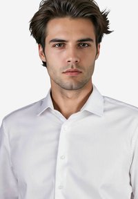 Junger Mann mit gestyltem Haar, der ein weißes Hemd mit Knöpfen trägt und direkt in die Kamera blickt, vor einem neutralen Hintergrund.