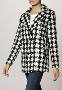 Schwarz-weißes Hahnentritt-Zweireiher-Mantel mit großen Knöpfen, Reverskragen und weicher Textur, getragen über einem weißen Oberteil und Jeans.