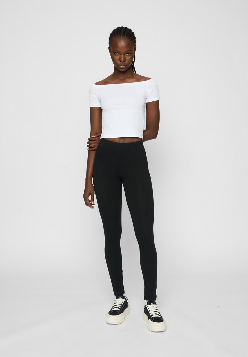 Zwarte stretchlegging gecombineerd met een witte off-shoulder crop top en zwarte sneakers. Glad materiaal met een slanke pasvorm.