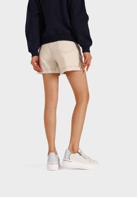 Sneakers bianchi con accenti argentati, abbinate a pantaloni corti beige. Il design presenta una texture liscia, punta rotonda e suole piatta.